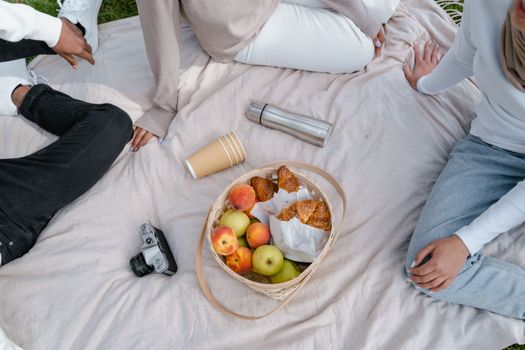 Pique-nique sur une couverture avec des fruits, des croissants et une bouteille, entouré de personnes en tenue décontractée.