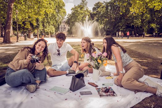 Groupe d'amis assis sur une couverture, prenant des photos et profitant d'un pique-nique dans un parc ensoleillé.