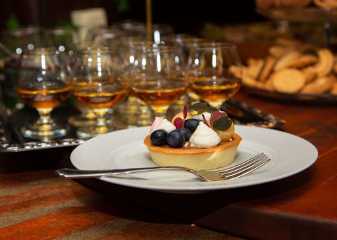 Tarte aux myrtilles et poires garnie de crème, servie avec des verres de liqueur en arrière-plan.